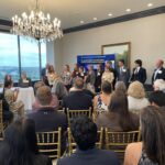 A large contingent of students, faculty and administrators from the South Carolina Governor's School of Science and Math stand in front of the display and room at the 2025 Meet the Finalists - Neval Erturk holds a microphone as she introduces her Finalist teams