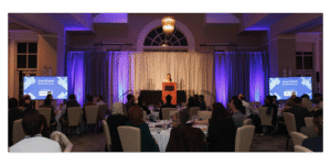 a view of the 2024 InnoVision Awards taken from the rear of the banquet room with attendees seated, a speaker at a podium at the center and display screens on either side of the stage, on which the speaker stands in the center