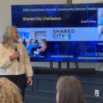 Kristin Parker of Shared City Charleston addresses attendees at Meet the Finalists - she looks over her shoulder at a screen displaying her headshot, along with her co-founder Holly Kelly, her logo, web address and photo of a Shared City event.